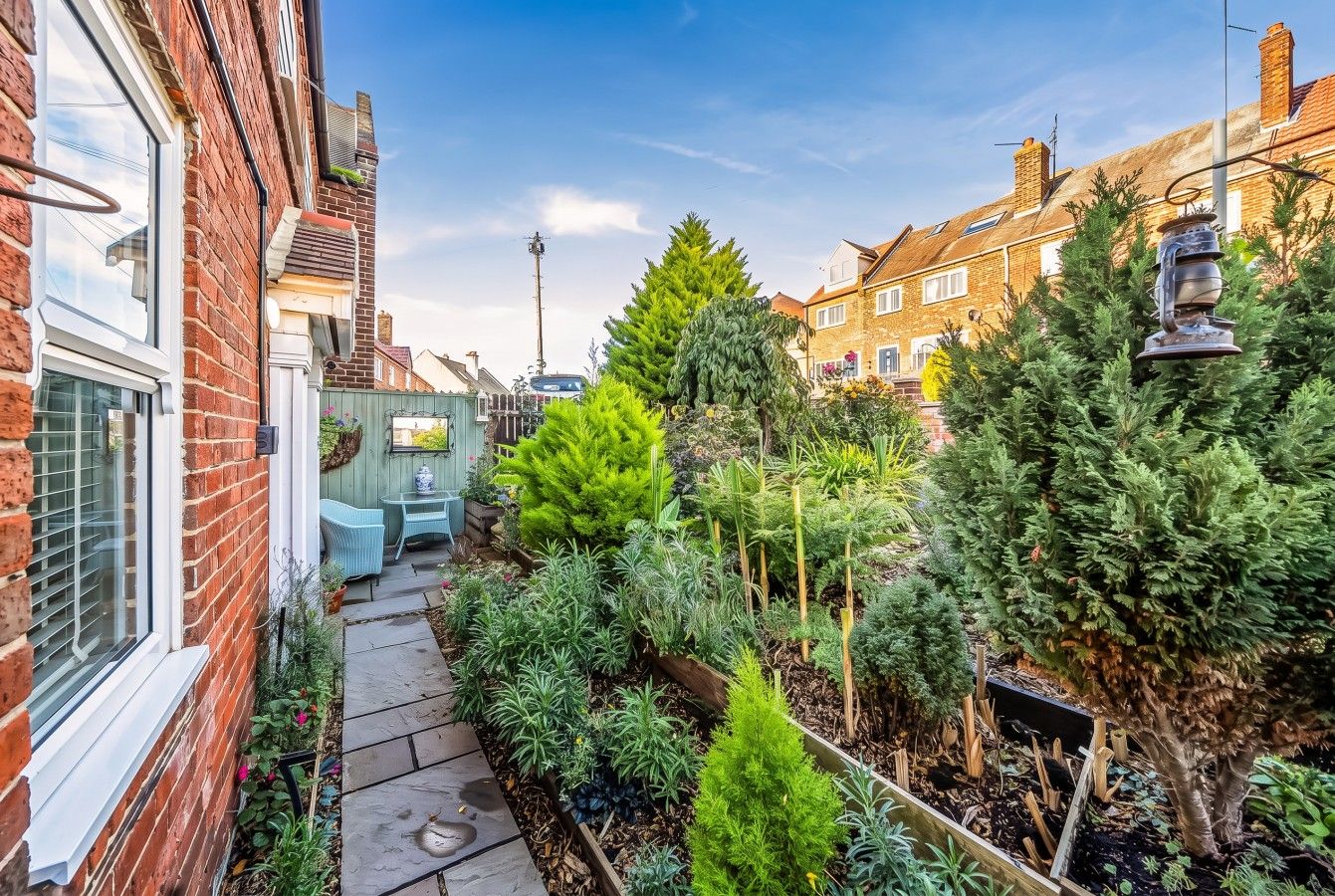 Un étroit sentier de jardin bordé de maisons en briques, agrémenté de diverses plantes en pot et d’un petit espace aménagé pour s’asseoir.