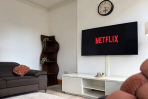 Un saló amb un televisor de pantalla plana fixat a la paret que mostra 'NETFLIX' en lletres vermelles, un sofà de color fosc amb un coixí texturat, un moble de televisió blanc i una estanteria de fusta amb objectes decoratius.