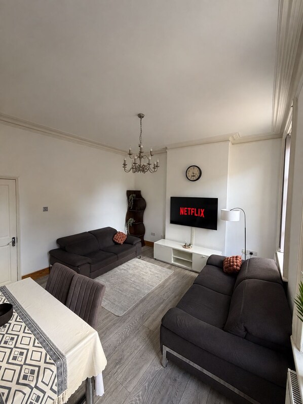 A living room with a sofa, a TV displaying 'NETFLIX,' a chandelier, and a clock on the wall.