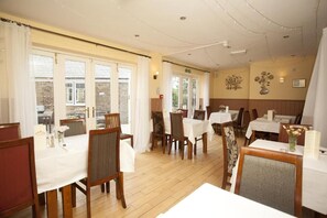 A dining area with multiple tables and chairs, a wooden floor, and a view through large windows. - The Five Bells (Ringwould)