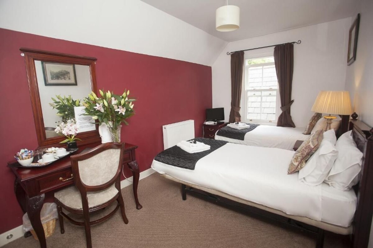 A hotel room with a bed, a desk with a chair, a lamp, a window with curtains, and a vase with flowers.