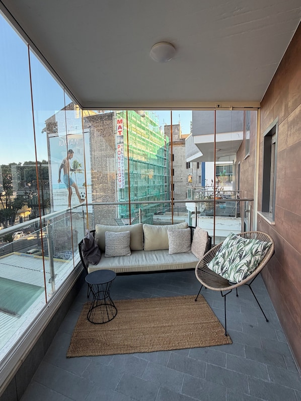 Varanda moderna com estrutura de vidro, um sofá, uma poltrona e vista para uma área de construção.