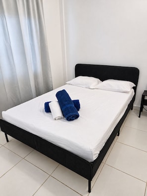 A single bed with white bedding and two rolled towels, one blue and one white, in a room with a black headboard and a small bedside table. - Hotel Canela (Punta Cana)