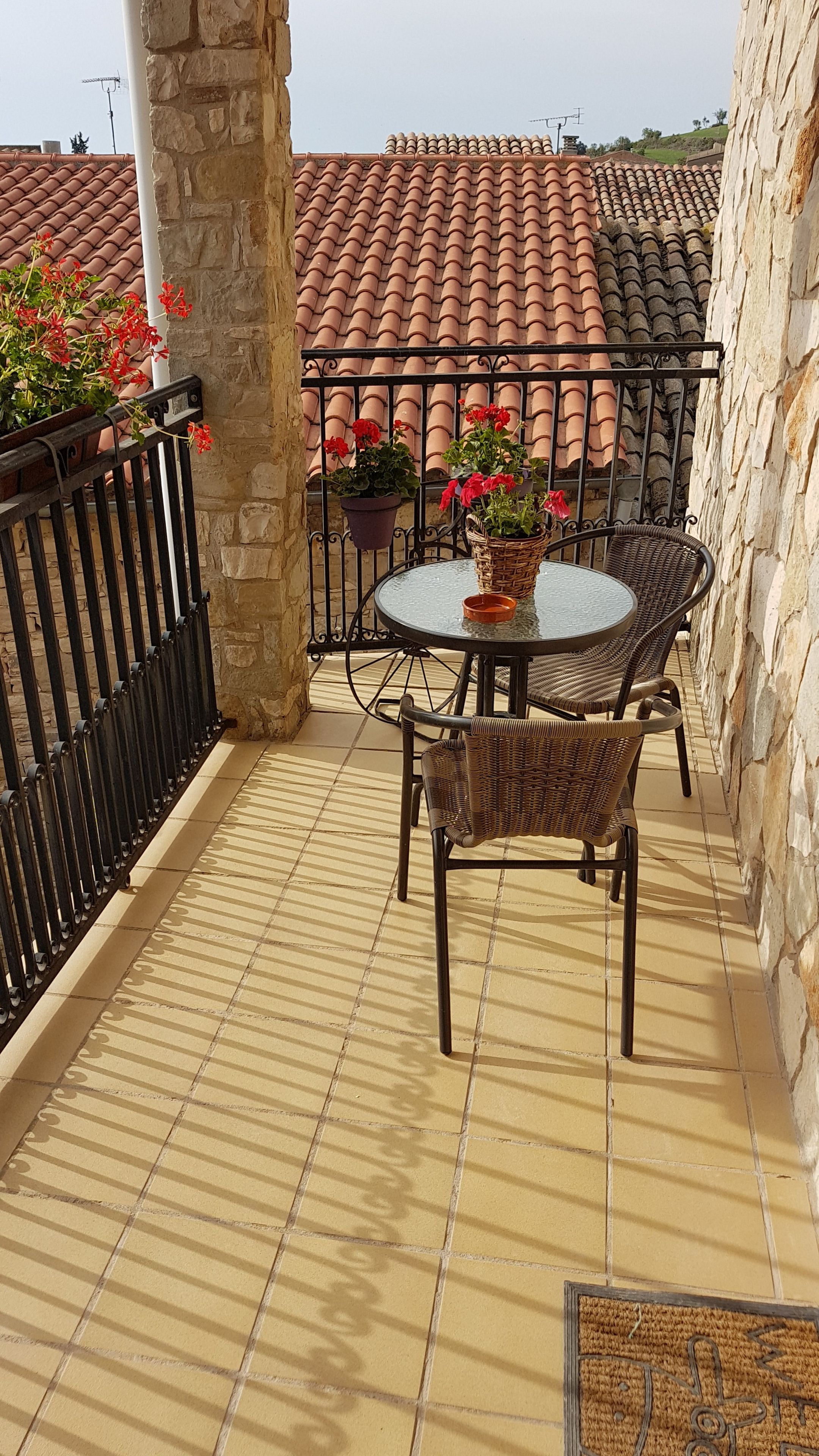 Un balcon avec une table ronde et des chaises en osier, des plantes en pot et un sol carrelé.