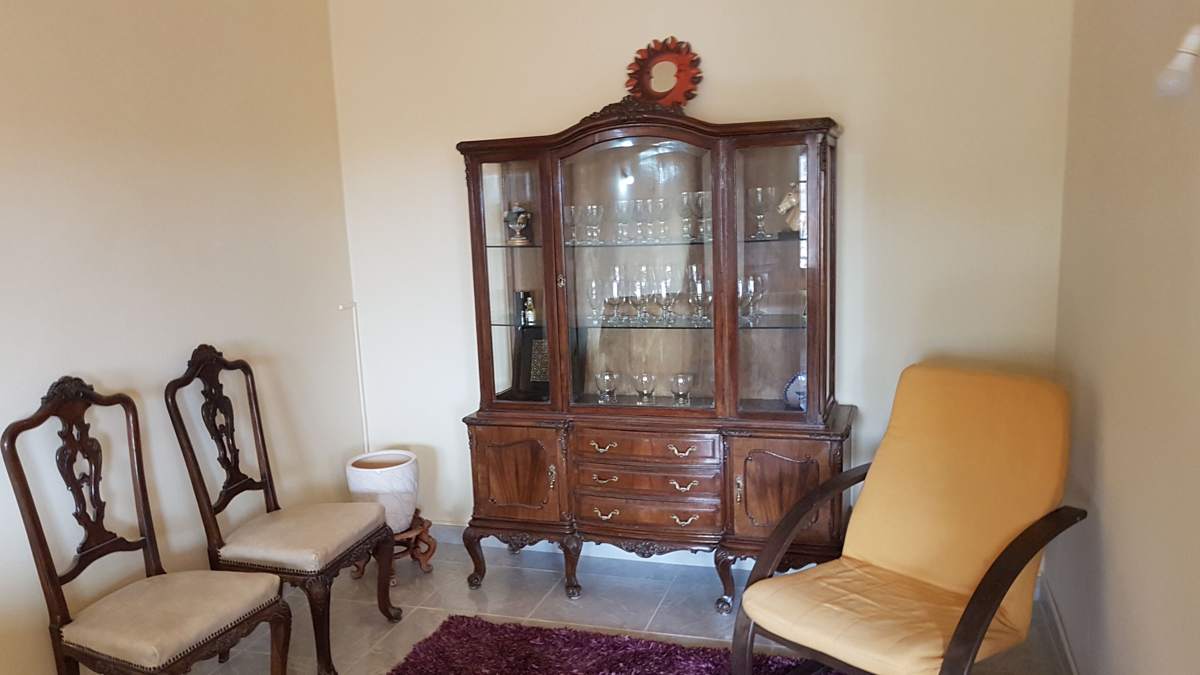 A room with a wooden cabinet, two chairs, a yellow armchair, and a purple rug.