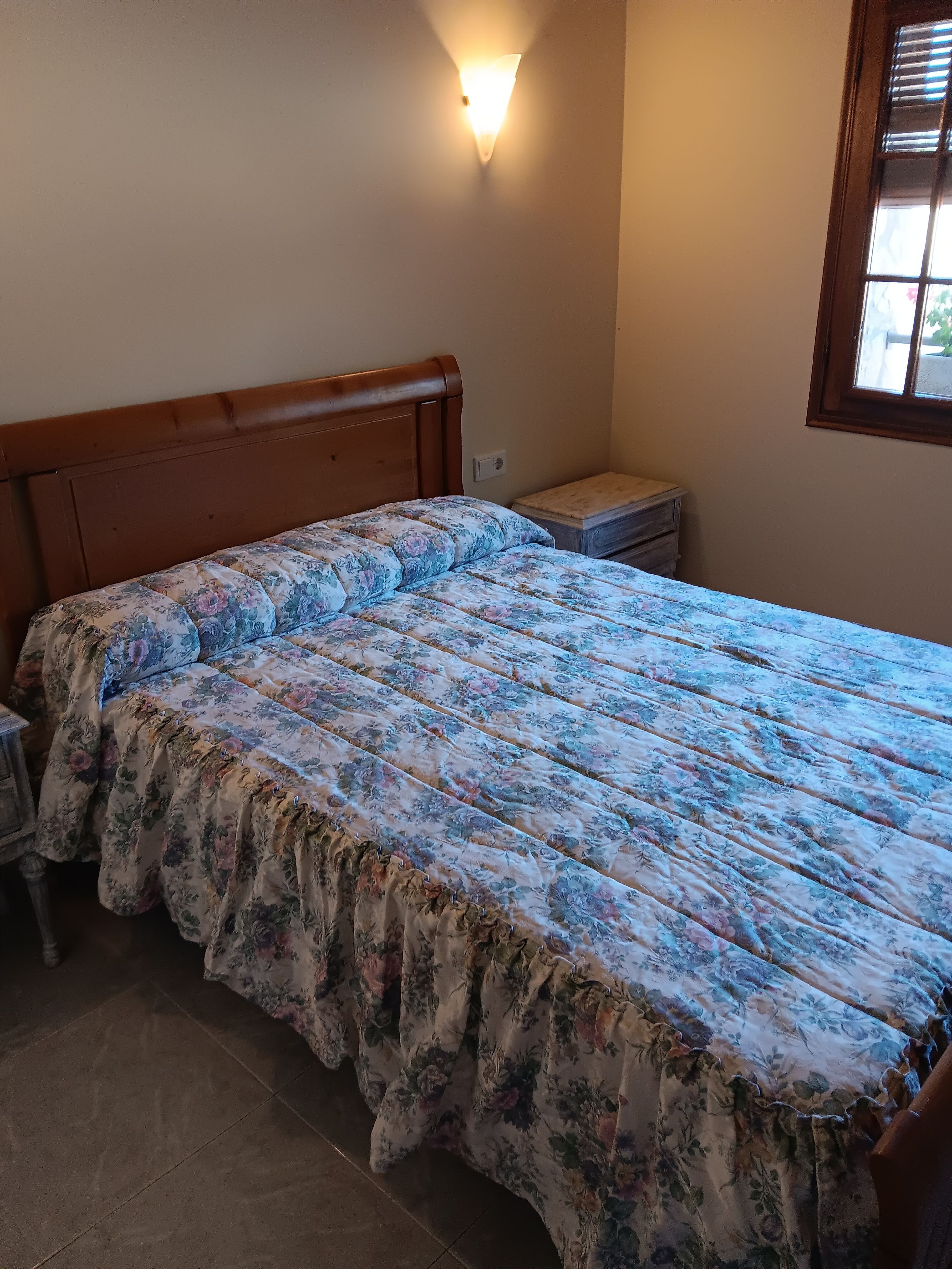 A bedroom with a wooden headboard, a floral bedspread, a bedside table, and a window with blinds.