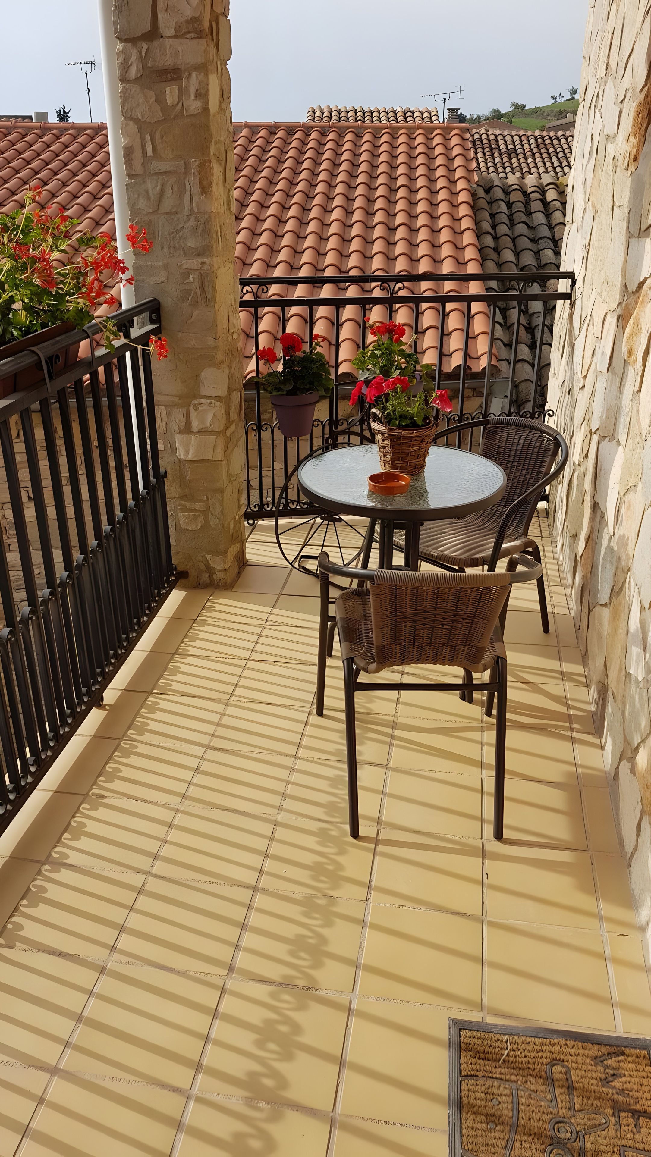 Een balkon met een ronde tafel, rieten stoelen, potplanten en een betegelde vloer.