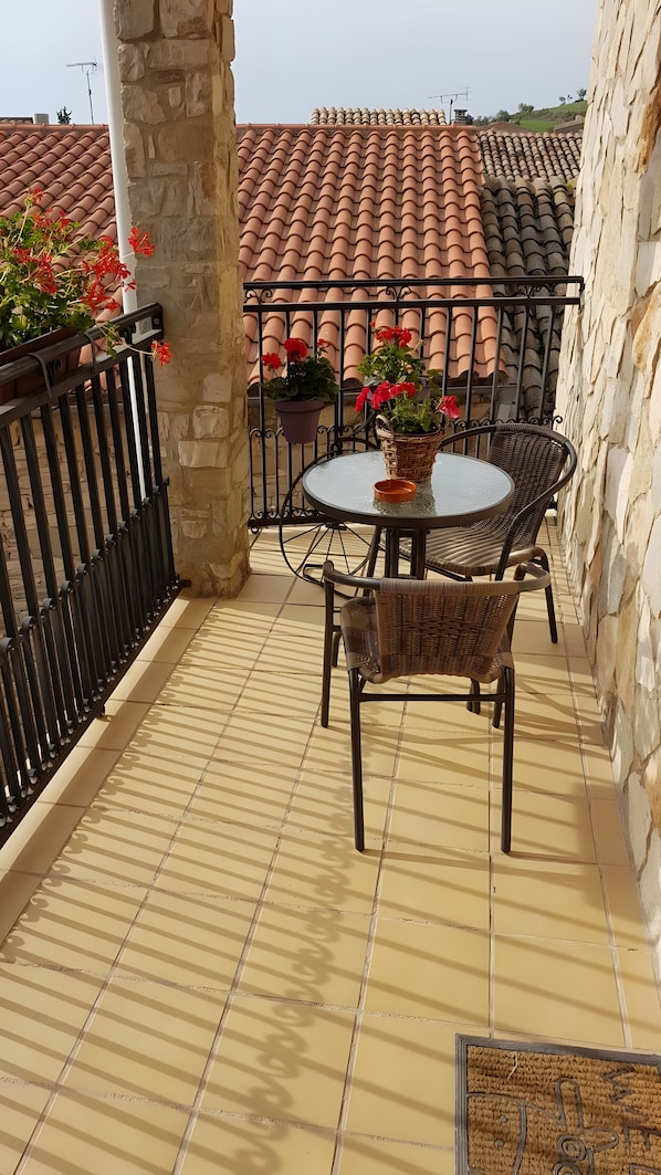 A balcony with a round table and wicker chairs, potted plants, and a tiled floor.