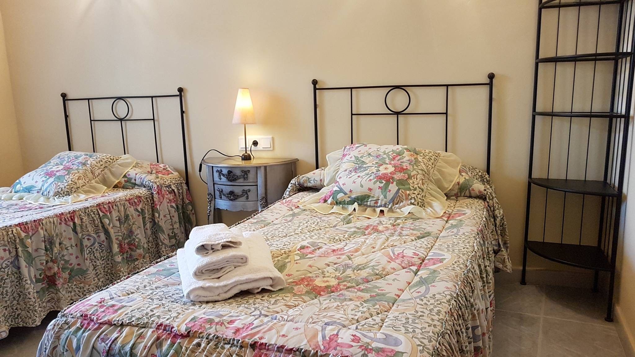 Una habitación con dos camas individuales, una mesita de noche con lámpara y una estantería metálica.