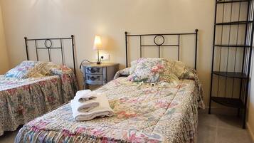 Una habitación con dos camas individuales, una mesita de noche con lámpara y una estantería metálica.