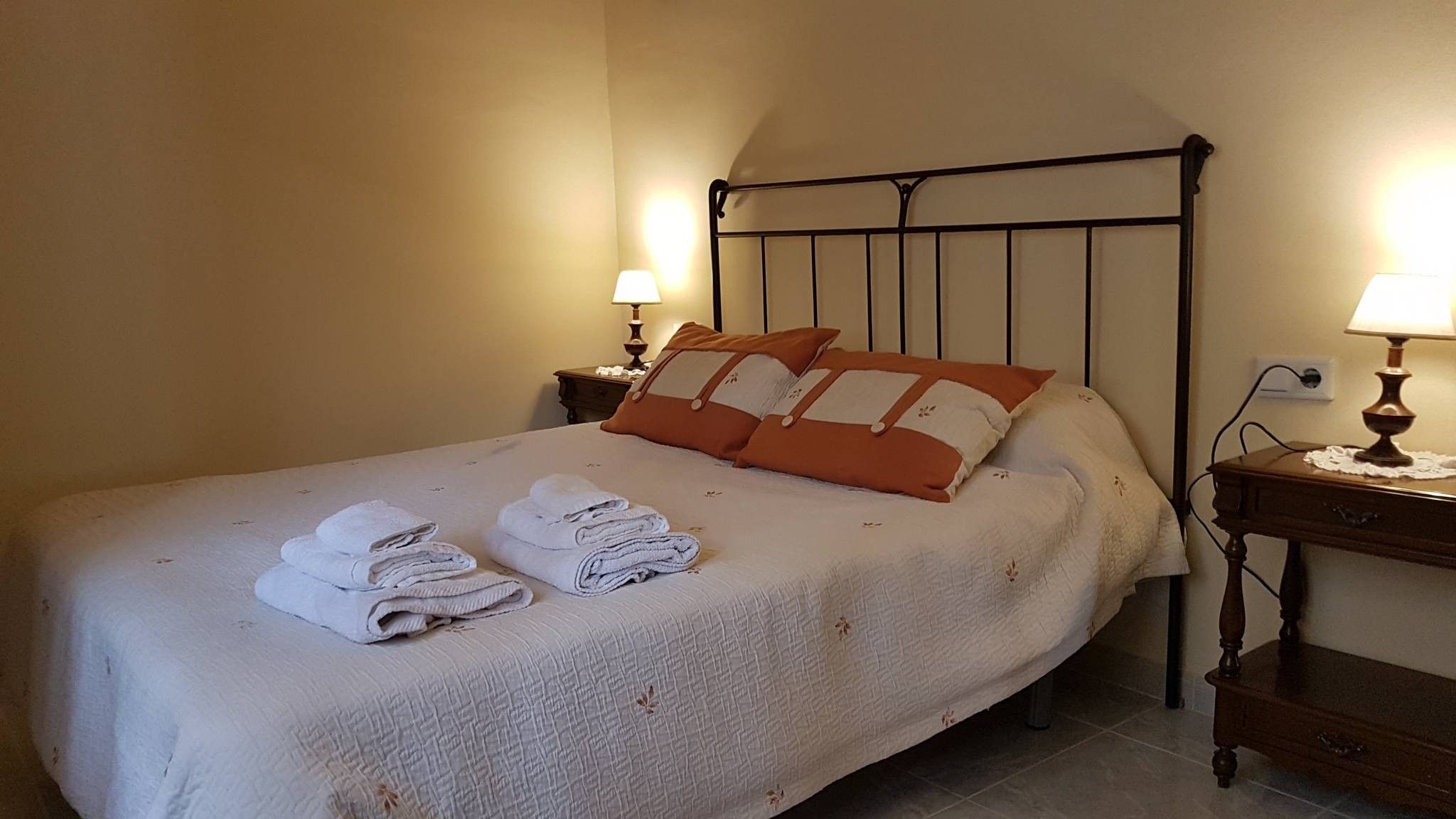 Een opgemaakt bed met opgevouwen handdoeken, een metalen hoofdbord, bedlampjes en een houten nachtkastje.