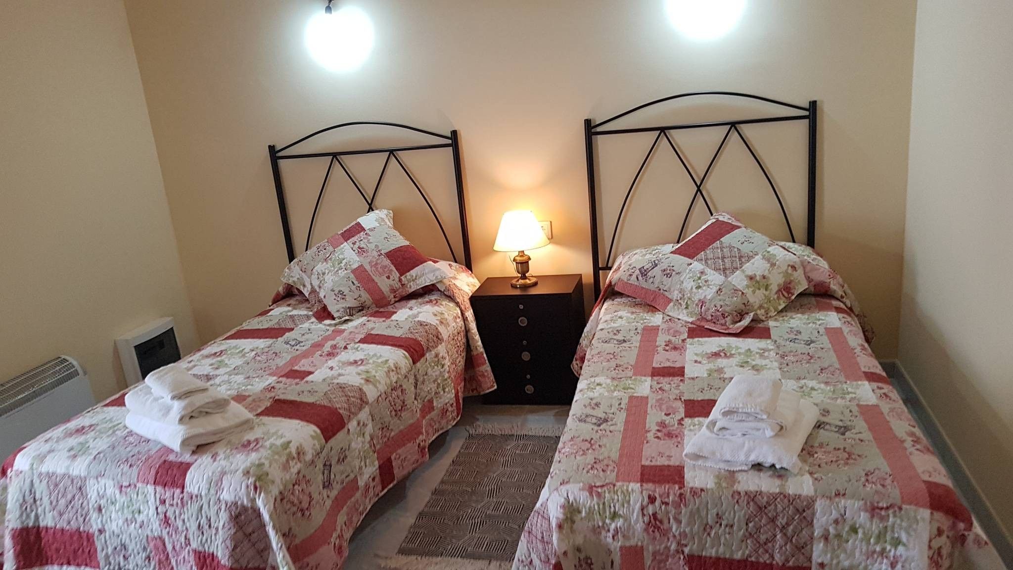 A room with two single beds, each with a floral-patterned bedspread and pillows, a nightstand with a lamp, and a radiator.