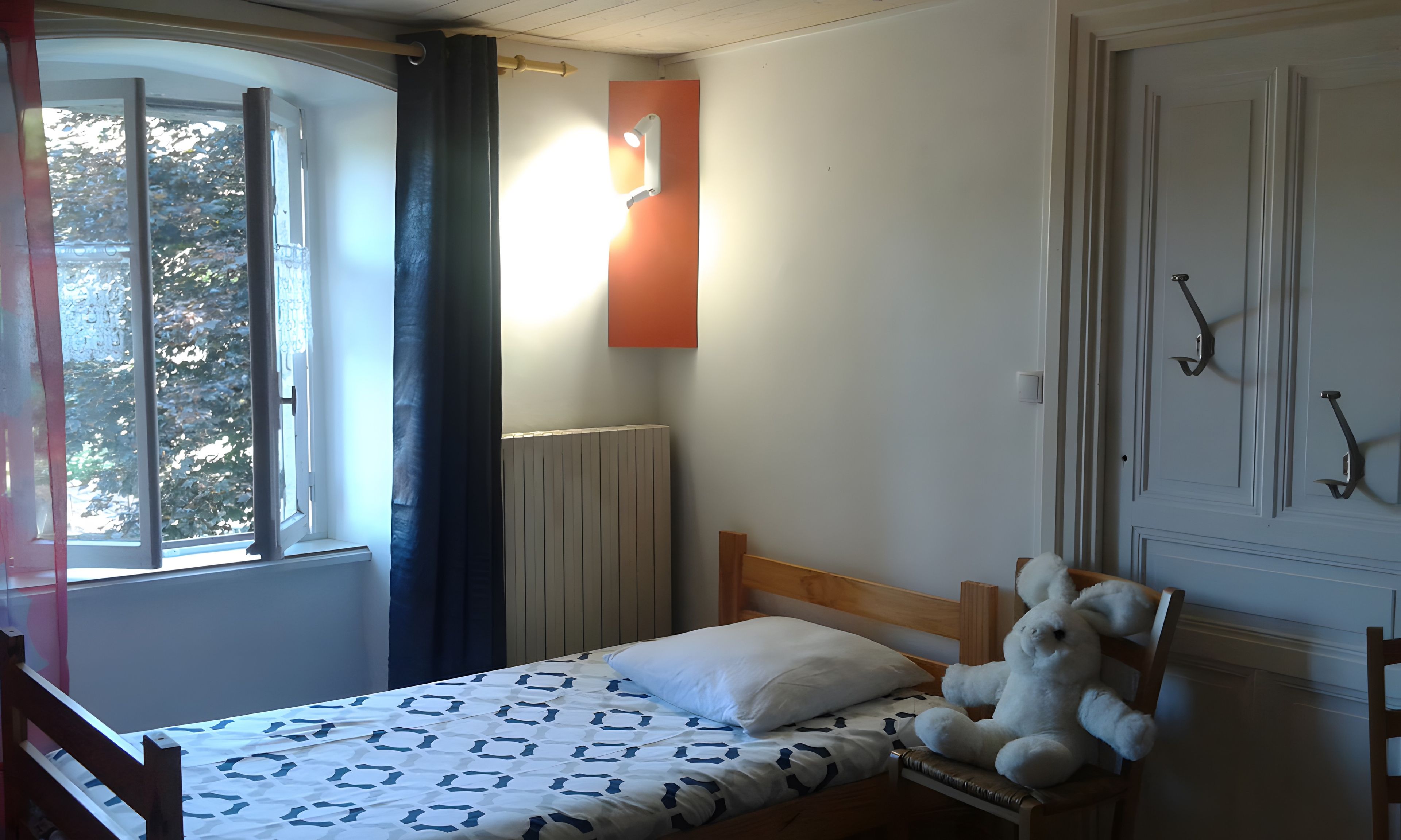 A bedroom with a bed, a window with curtains, a radiator, and a stuffed toy on the bed.