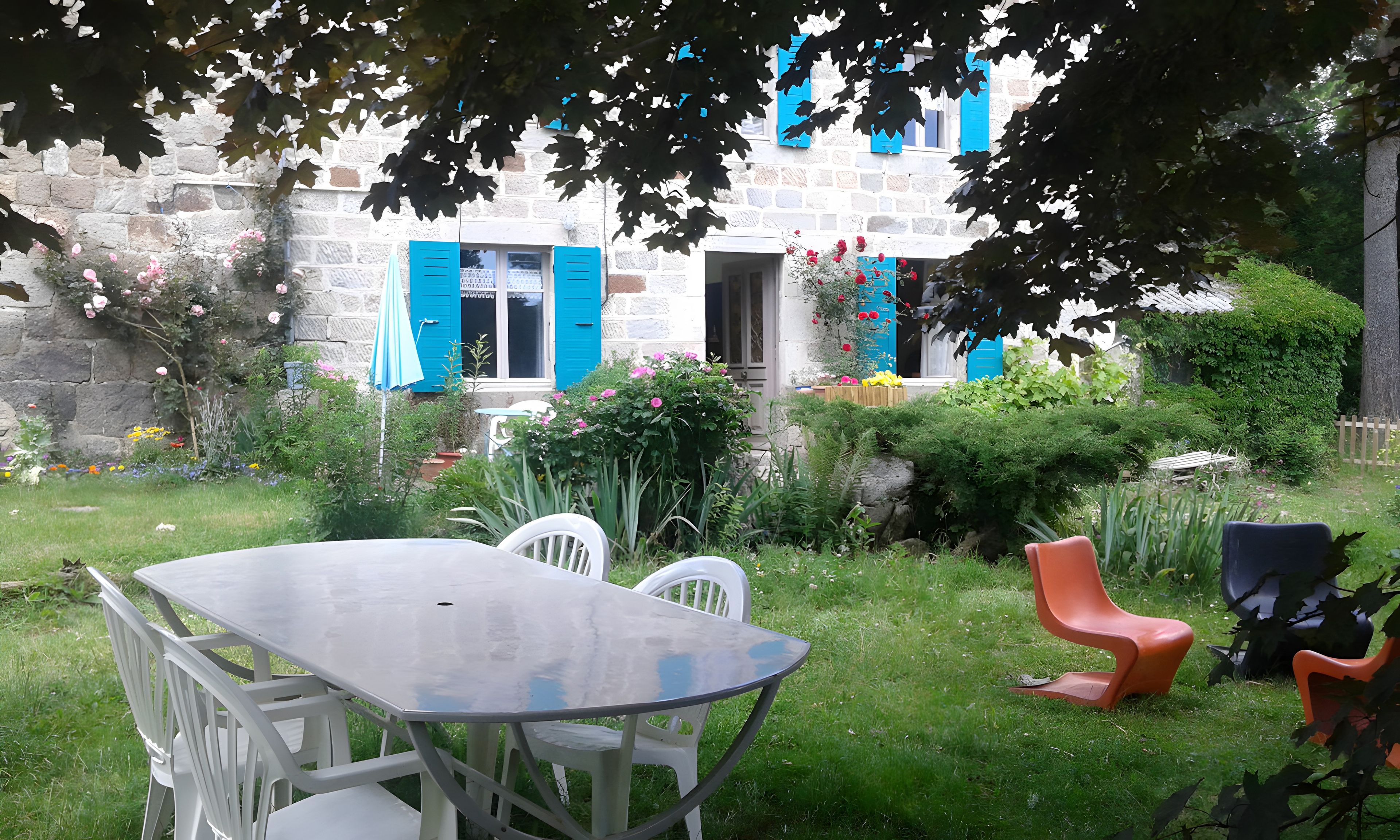 Un jardin avec une table et des chaises, un bâtiment en pierre avec des volets bleus, et une variété de plantes et de fleurs.