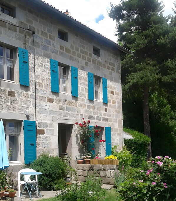 A stone building with blue shutters, a garden with potted plants, and a white chair.