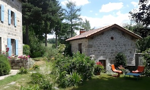 Une maison en pierre avec un toit en tuiles, entourée d’un jardin où poussent différentes plantes et fleurs.