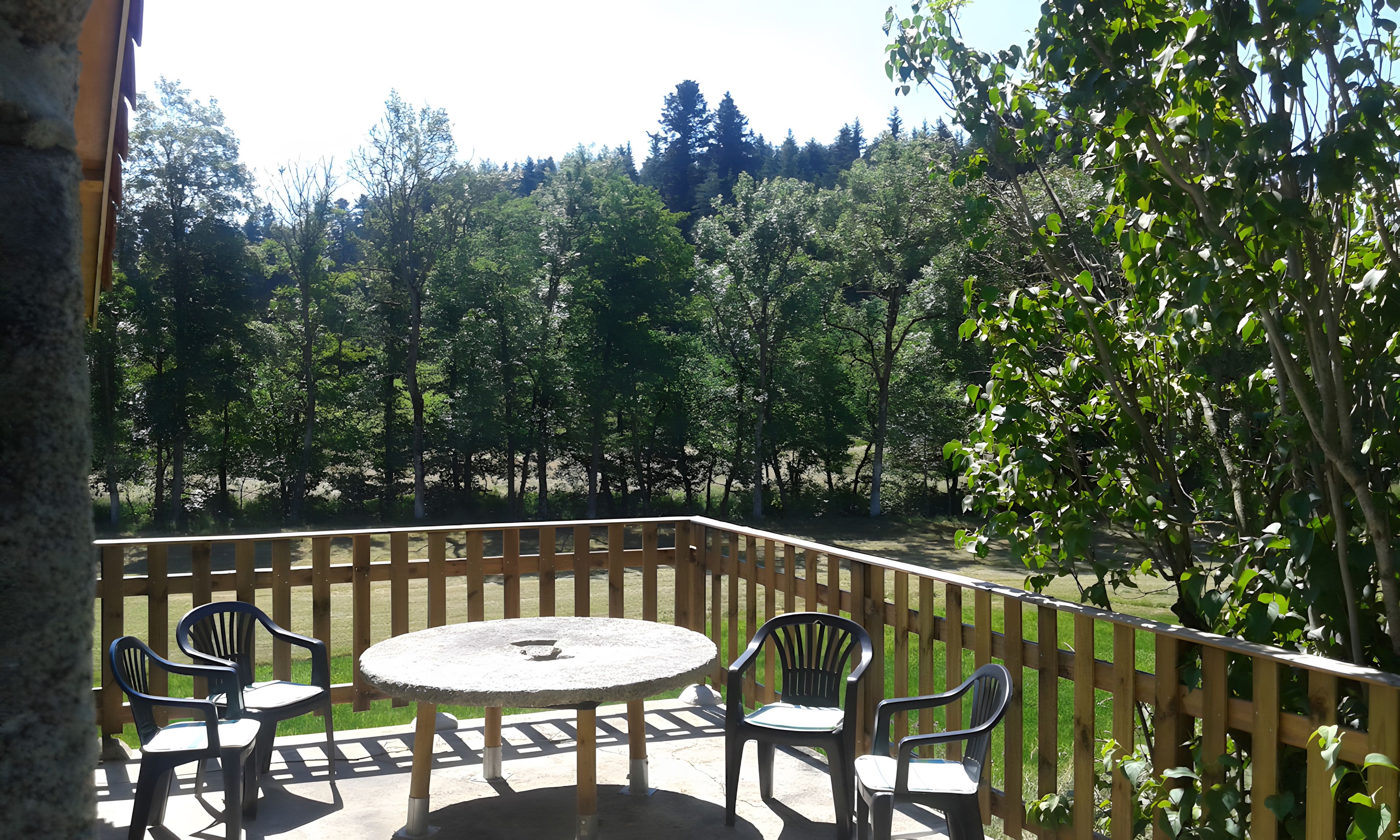 A patio with a round table and four chairs, surrounded by a wooden fence, overlooking a forested area.