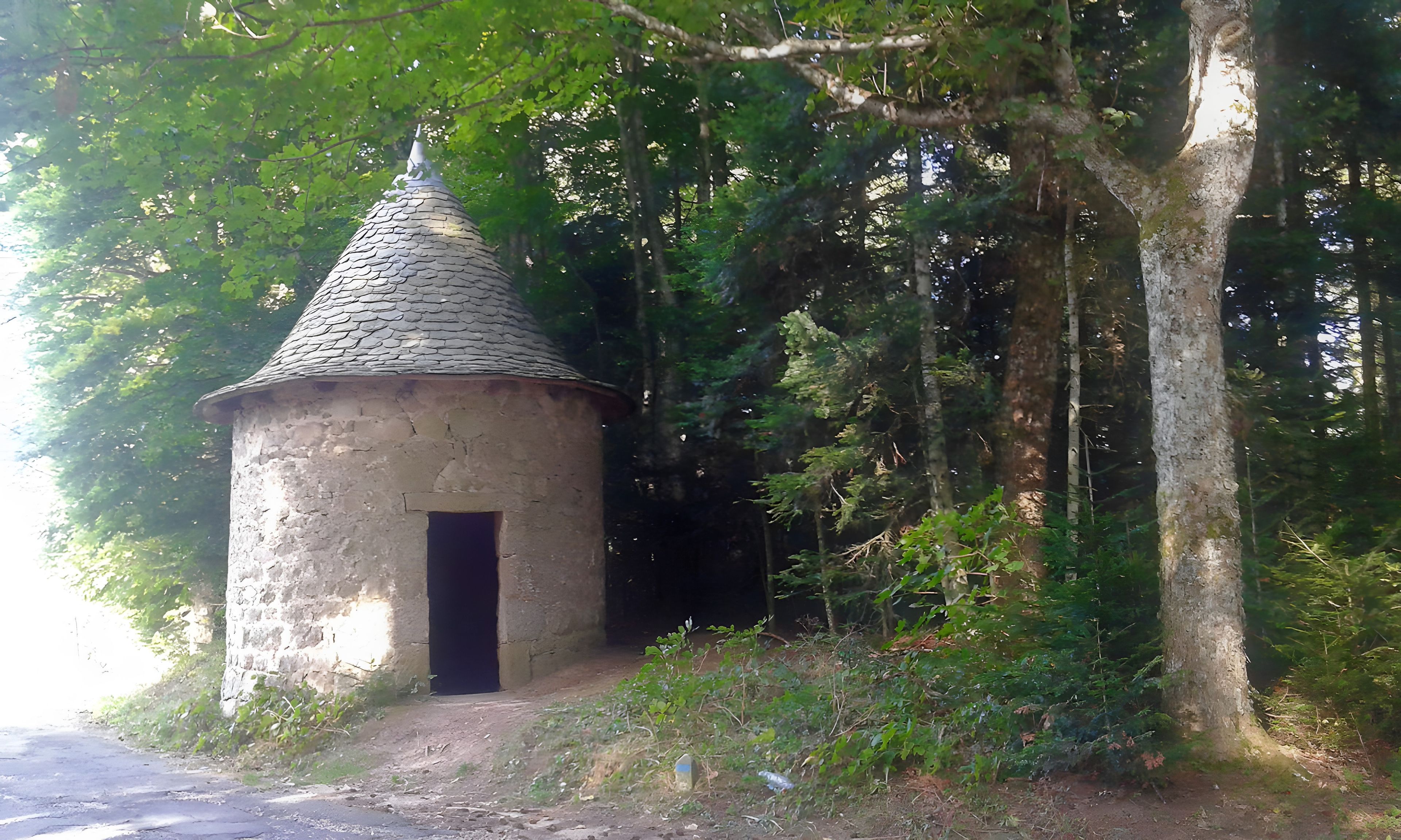 A small stone structure with a conical roof and a dark entrance, surrounded by dense forest.