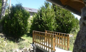 A wooden deck with a stone wall, surrounded by dense green foliage and a house in the background. - Cottage 'Le Petit Moulin' (Chenereilles)