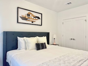 A neatly made bed with a dark headboard and white bedding, a framed abstract artwork above it, a bedside lamp, and a white door to the right. - Stylish 1B1B with Modern Finishes (Atlanta)