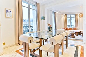 A modern living room with a glass dining table, white upholstered chairs, a sofa with orange cushions, and a view of the city through large windows. - Parisian Luxury with Eiffel Tower Views & Designer Interiors (Paris)