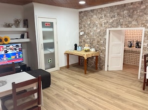 A room with a dining table, chairs, a television, a door with an emergency sign, a stone wall, and a shelf with bottles.