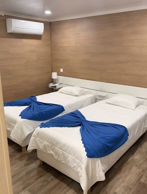 A hotel room with two beds, a bedside table with a lamp, and an air conditioning unit on the wall.