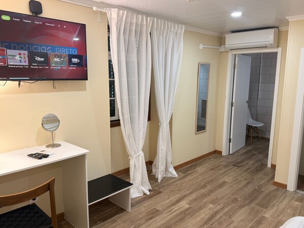 Een hotelkamer met een televisie waarop een nieuwskanaal wordt vertoond, een bureau met een spiegel, een stoel en een gordijn.