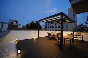 Une terrasse extérieure avec un plancher en bois, une pergola dotée d’un éclairage et une table avec des chaises.