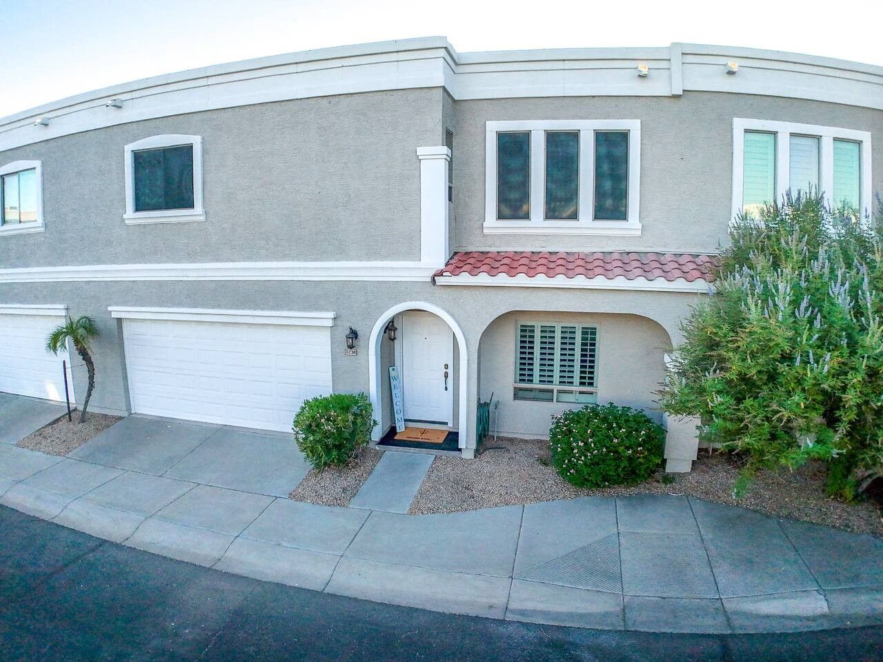 Una casa de dos pisos con garaje, una puerta principal con felpudo y un pequeño jardín delantero con plantas.