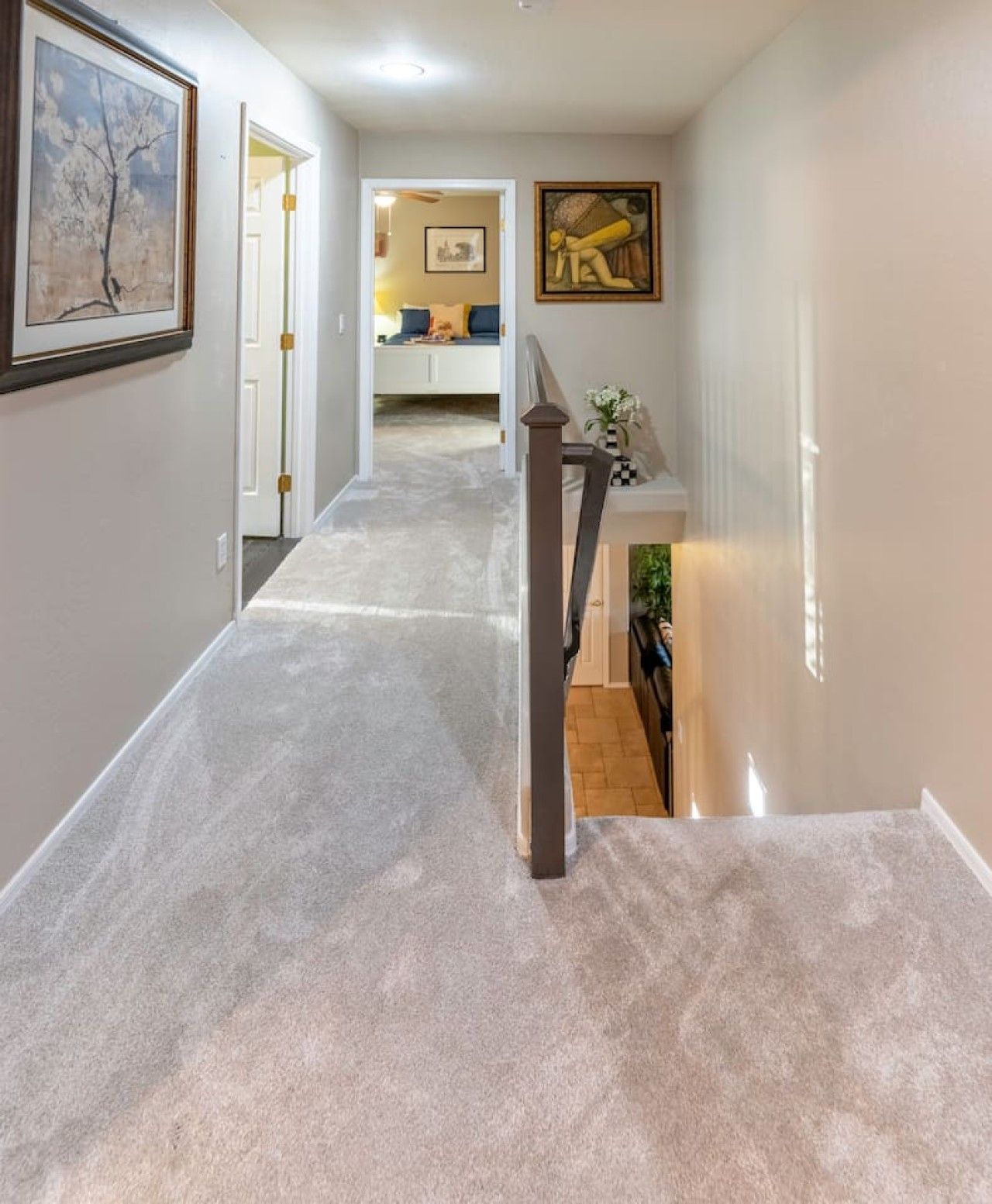Un pasillo con suelo alfombrado, una escalera con barandilla y una puerta que da a una habitación con cama y un cuadro en la pared.