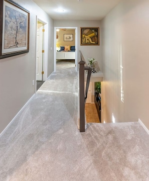 Un pasillo con suelo alfombrado, una escalera con barandilla y una puerta que da a una habitación con cama y un cuadro en la pared.