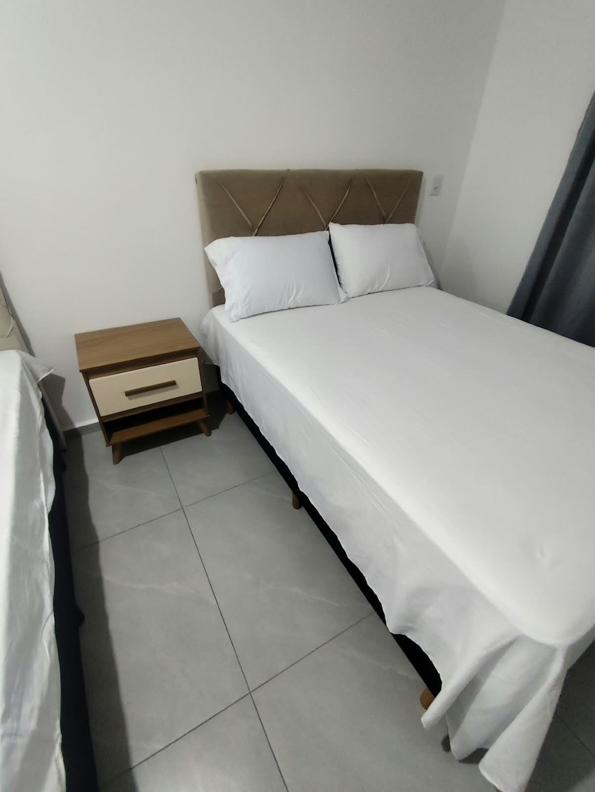 A single bed with a wooden headboard and two pillows, a wooden nightstand with a drawer, and a tiled floor.