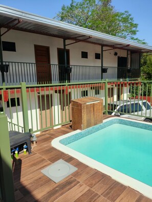 Una piscina con bordo piastrellato, un patio coperto con tetto in metallo e un recinto verde che circonda la proprietà.