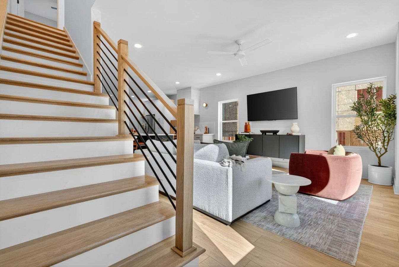 A modern living room with a wooden staircase, a grey sofa, a red accent chair, and a flat-screen TV mounted on the wall.