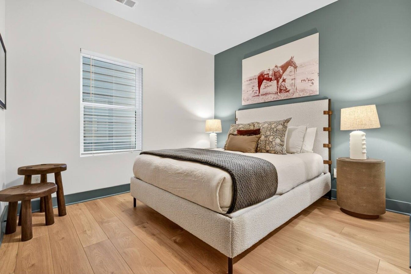 A bedroom with a bed, two bedside tables, a window with blinds, and a painting on the wall.