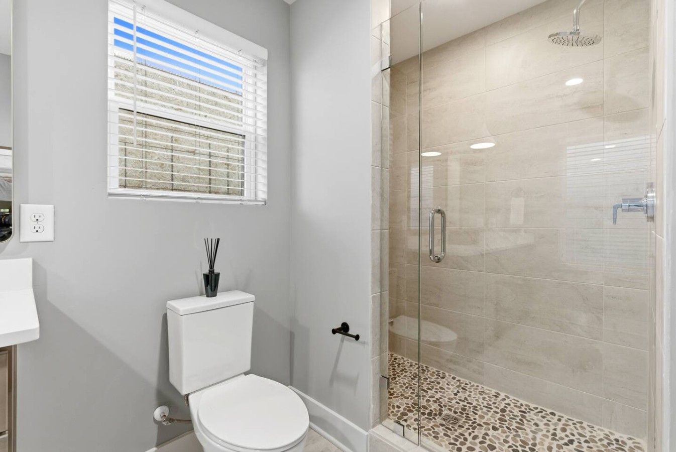 A bathroom with a toilet, a glass-enclosed shower, and a window with blinds.