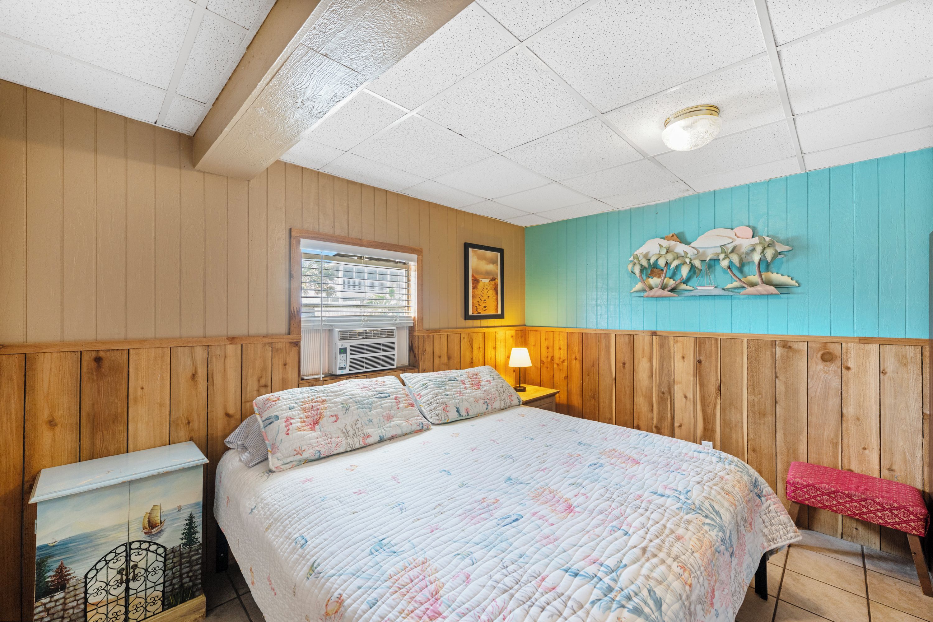 A bedroom with a wooden paneled wall, a bed with floral bedding, a nightstand, a lamp, and a wall mural.