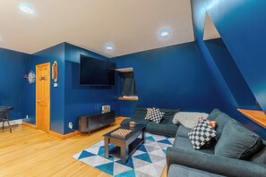 Ein modernes Wohnzimmer mit blauer Akzentwand, einer Holztür, einem Flachbildfernseher, einer Couch mit gemusterten Kissen und einem Couchtisch mit Schachbrett.
