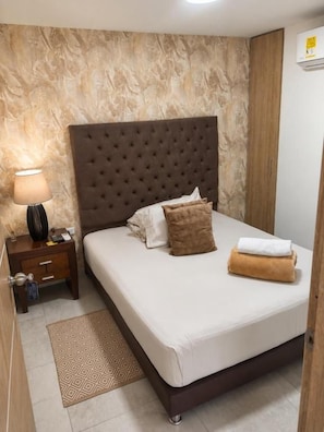 A neatly made bed with a tufted headboard, a bedside table with a lamp, and a patterned rug. - HOTEL BELEN La Flora  (Cali)