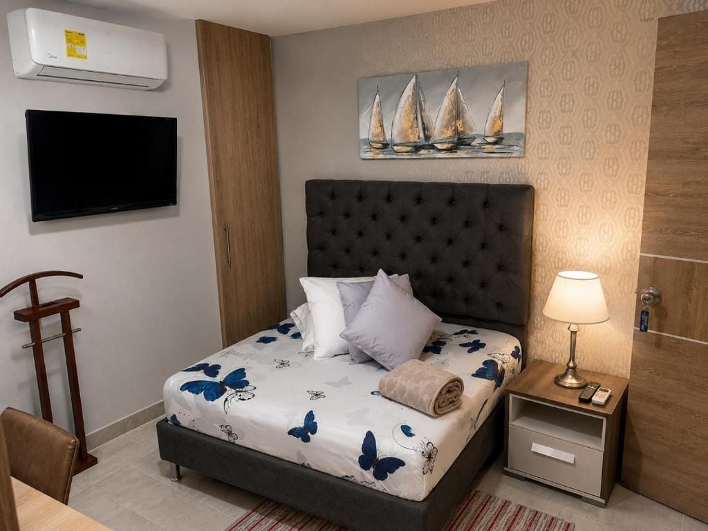 A hotel room with a bed, a TV, a painting, a bedside table with a lamp, and a wooden door.