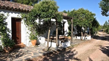 Un bâtiment rustique avec un toit en tuiles, une porte en bois et un chemin de pierre menant à un espace extérieur abrité, aménagé pour s’y asseoir.