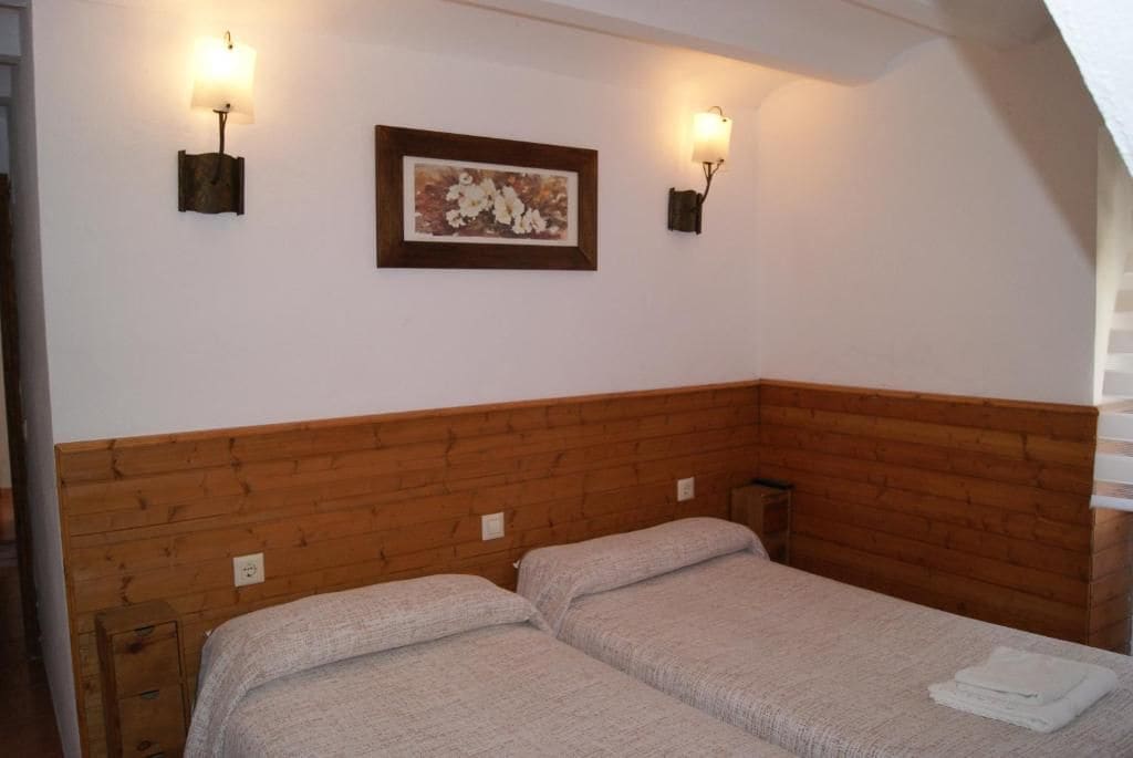 A room with two beds, wooden headboard, wall-mounted light fixtures, and a framed picture.