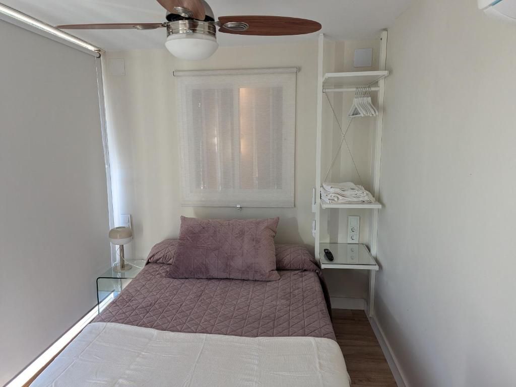 Un dormitorio con cama, ventilador de techo, ventana con cortinas, mesita de noche con lámpara y estante empotrado con ropa.