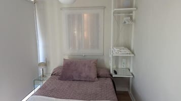 Un dormitorio con cama, ventilador de techo, ventana con cortinas, mesita de noche con lámpara y estante empotrado con ropa.