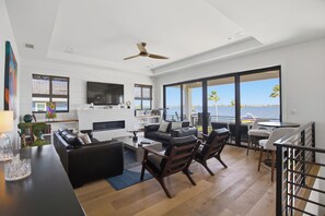A spacious living area with a large window overlooking a body of water, a kitchen with a flat-screen TV, and a seating area with a coffee table. - Manatee Bay Oasis- Waterfront Luxury + Heated Pool (Bradenton)