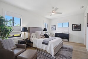 Ein Schlafzimmer mit einem Bett, Nachttischen, einem Schreibtisch, einem Stuhl, einem Deckenventilator und einem Fenster mit einer Pflanze.