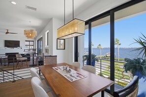 Uma sala de jantar moderna com uma mesa de madeira, cadeiras brancas e vista para o mar através de grandes janelas.
