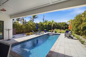 Piscina com espreguiçadeiras e guarda-sóis, rodeada por plantas tropicais e palmeiras.