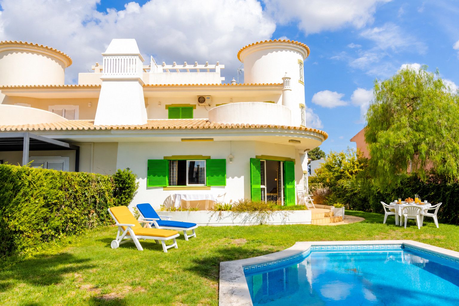 Une maison à deux étages avec une piscine, des volets verts et un espace salon extérieur.
